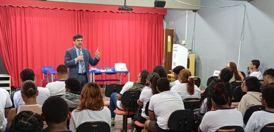 Alunos sentados assistem palestra feita por homem de terno, que aponta para duas urnas eletrônic...
