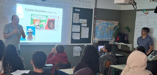 Alunos sentados assistem palestra feita por homem de terno, que aponta para duas urnas eletrônic...