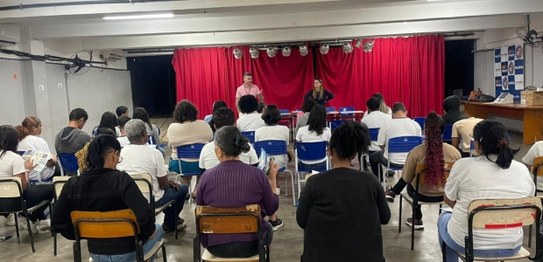 Auditório com cortina vermelha ao fundo, onde um homem está à frente, conduzindo uma apresentaçã...