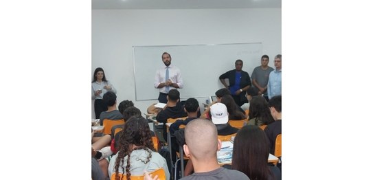 Alunos sentados assistem palestra feita por homem de terno, que aponta para duas urnas eletrônic...