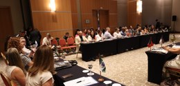 Foto do salão de convenção; uma mesa ao centro e duas fileiras de mesas dispostas à lateral, com...
