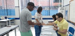 Foto de uma seção eleitoral durante o processo de votação. Um homem entrega seu documento a um m...