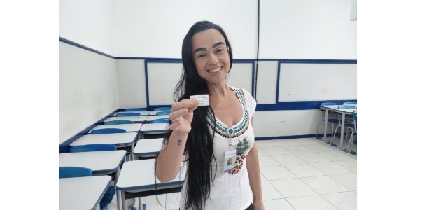 Eleitora posa segurando o comprovante de votação, dentro de uma seção eleitoral; ela tem cabelos...