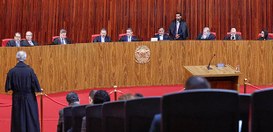 Magistrados vestindo togas pretas estão sentados lado a lado em uma longa e curva bancada de mad...
