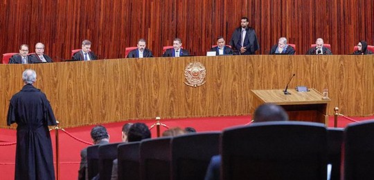 Magistrados vestindo togas pretas estão sentados lado a lado em uma longa e curva bancada de mad...
